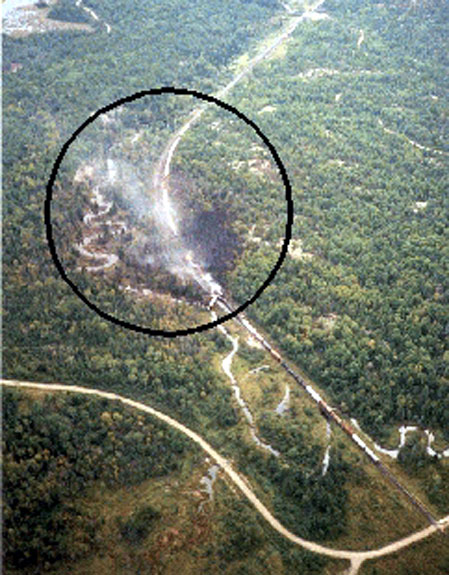 Aerial photograph of derailment site from the northwest. The circle denotes blackened area from fire damage. Note the public crossing at the bottom right. Note the direction of smoke and fumes, and the location of Little Key River adjacent to the right-of-way.