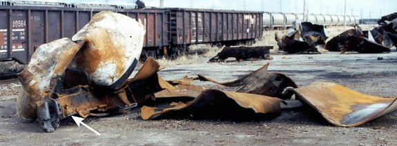 Main part of the B end of the car, including the head and approximately 16.5 feet of the flattened sections of the tank shell recovered after the derailment. (The coupler and stub sill are in the foreground at the lower left). The remaining pieces of the tank car and jacket can be seen in the right background.