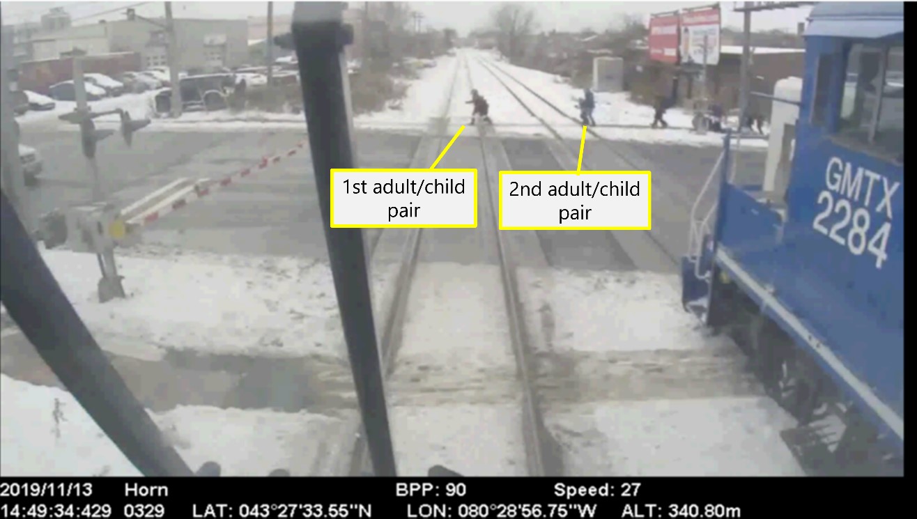 An image from the forward-facing video camera from GO 3919 train’s cab car 329 showing the 1st adult and child pair traversing the Lancaster Street West crossing followed by the 2nd adult and child pair (Source: GO Transit with TSB annotations)